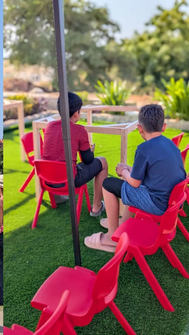 Dos niños sentados en sillas rojas frente a mesas de madera bajo el toldo